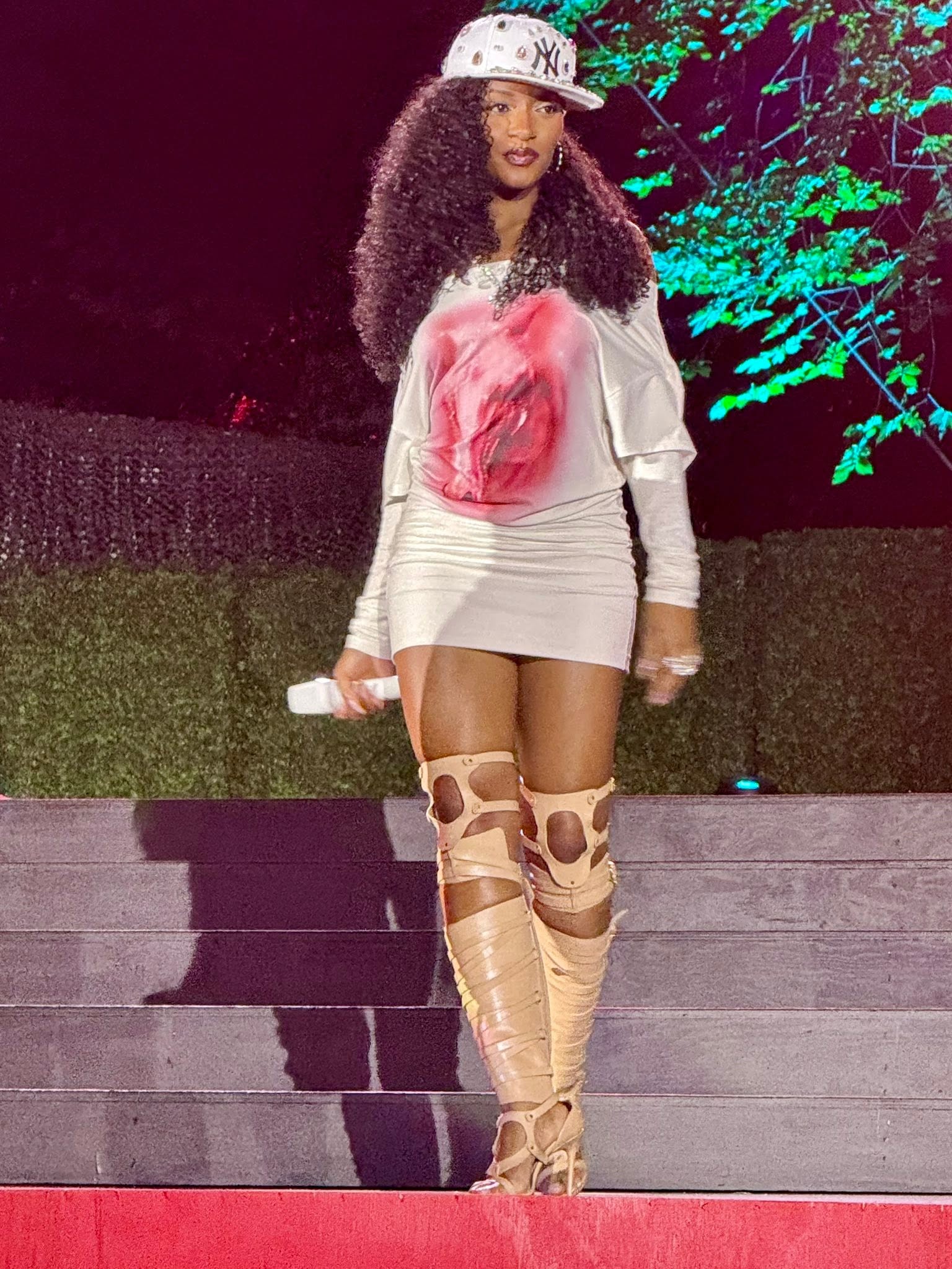 Ayra Starr performs onstage at night at the Global Citizen Festival: long curly hair, a white New York Yankees cap set with colorful rhinestones, an all-white outfit, and a microphone in her hand.