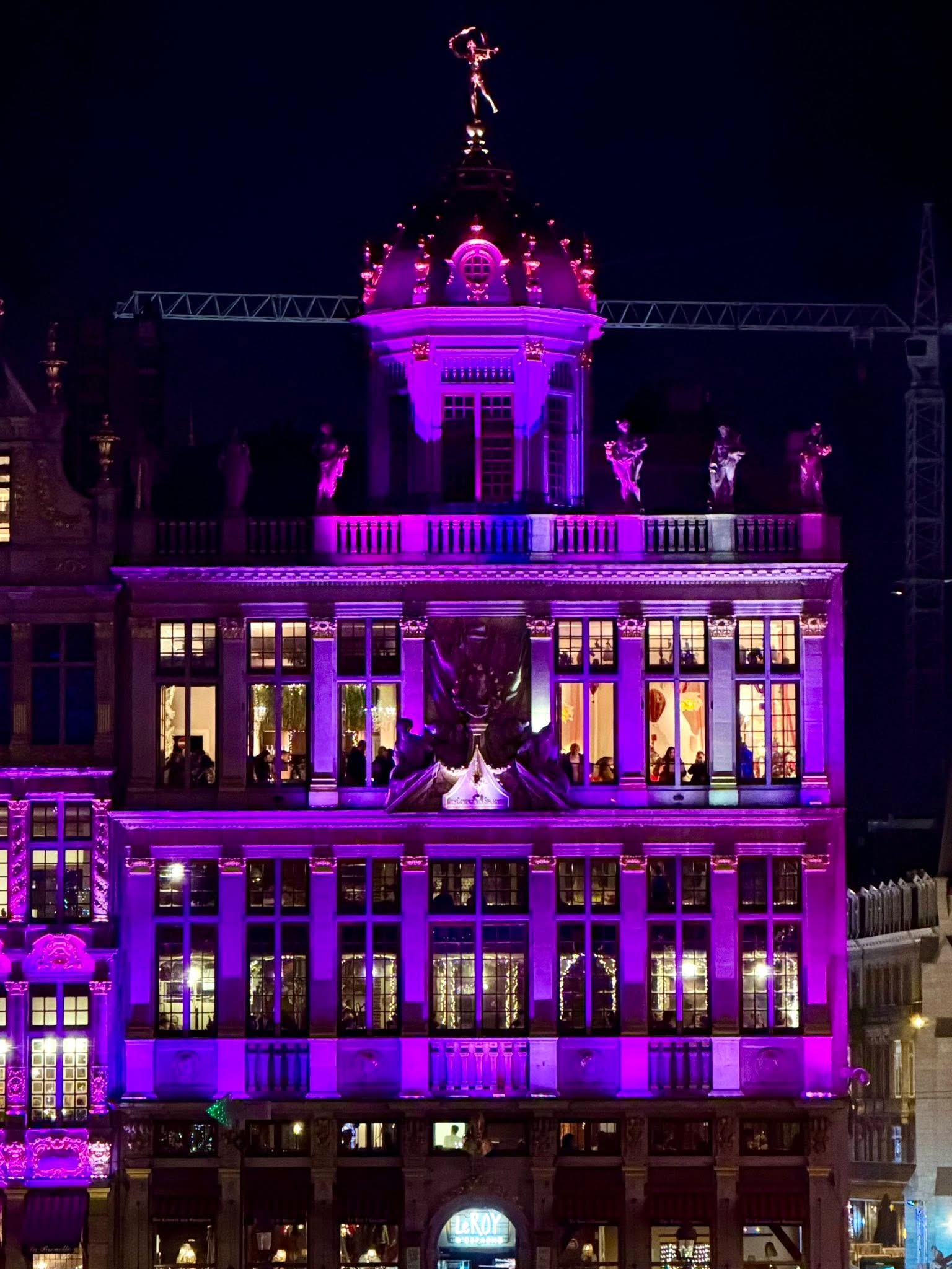 A guildhall on Brussels' Grand Place illuminated in violet light at night, a crane visible in the dark sky.
