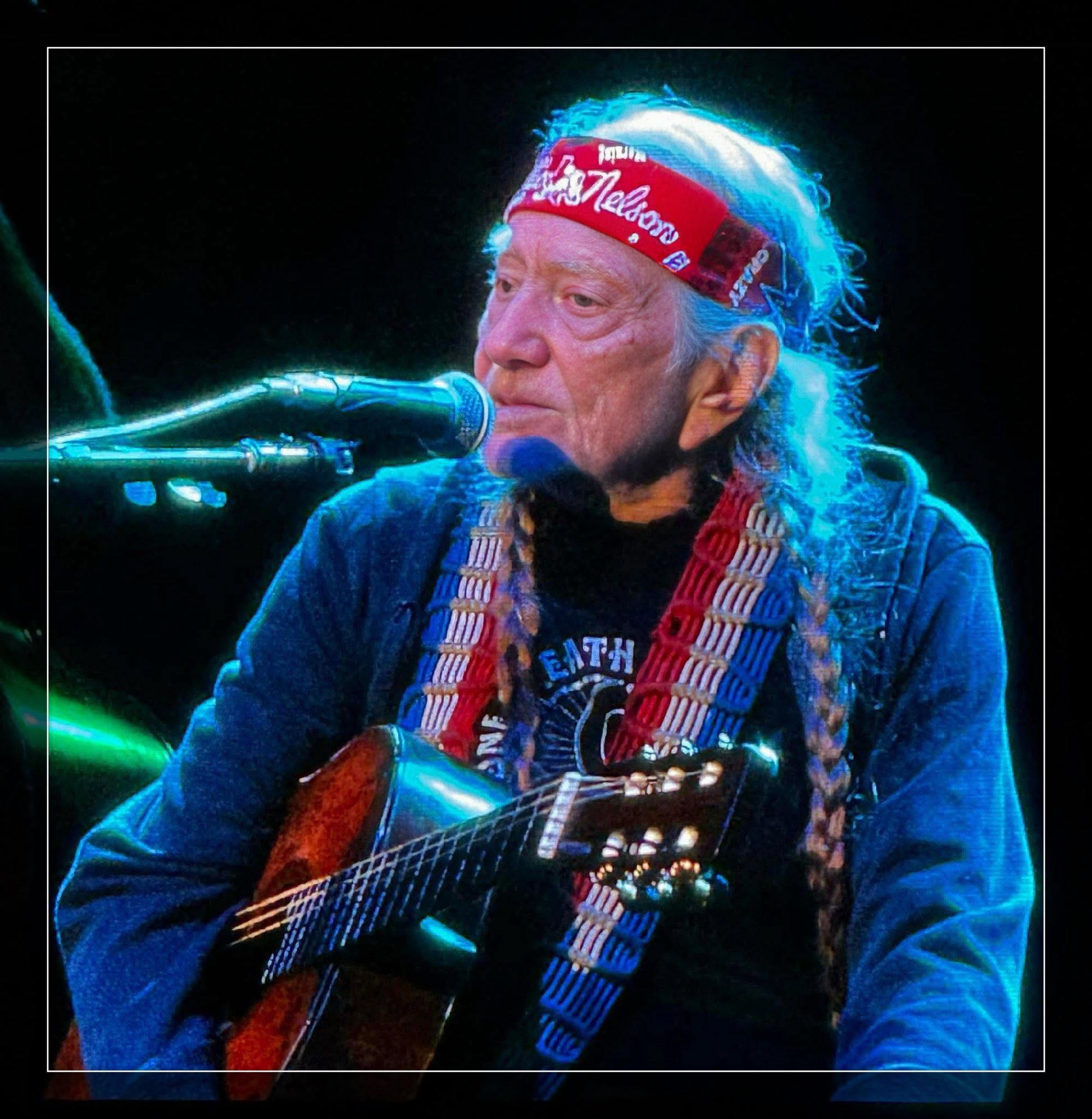 Willie Nelson performs onstage with his guitar, wearing his signature red bandana.