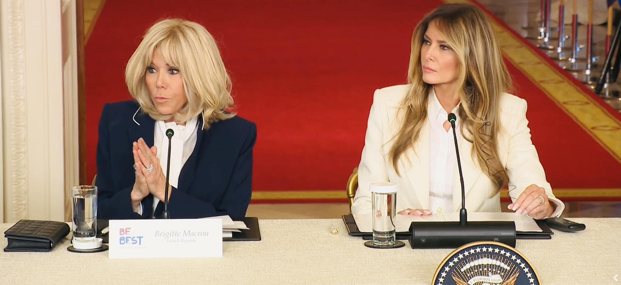 A video still of Brigitte Macron and Melania Trump seated at a table with microphones during a White House event; a 'BE BEST' nameplate sits before Mrs. Macron.