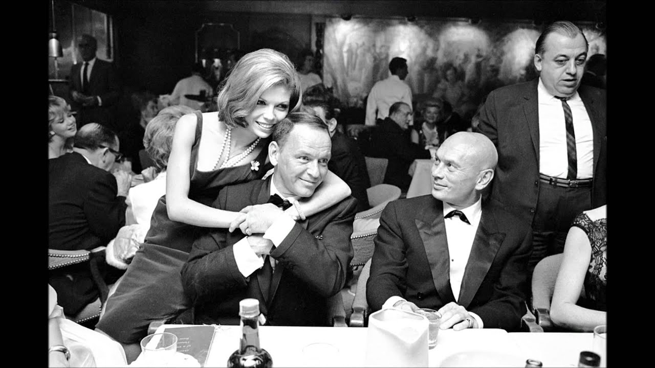 Nancy Sinatra embraces her father Frank Sinatra at a nightclub table, Yul Brynner seated beside them, in a vintage black-and-white photograph.