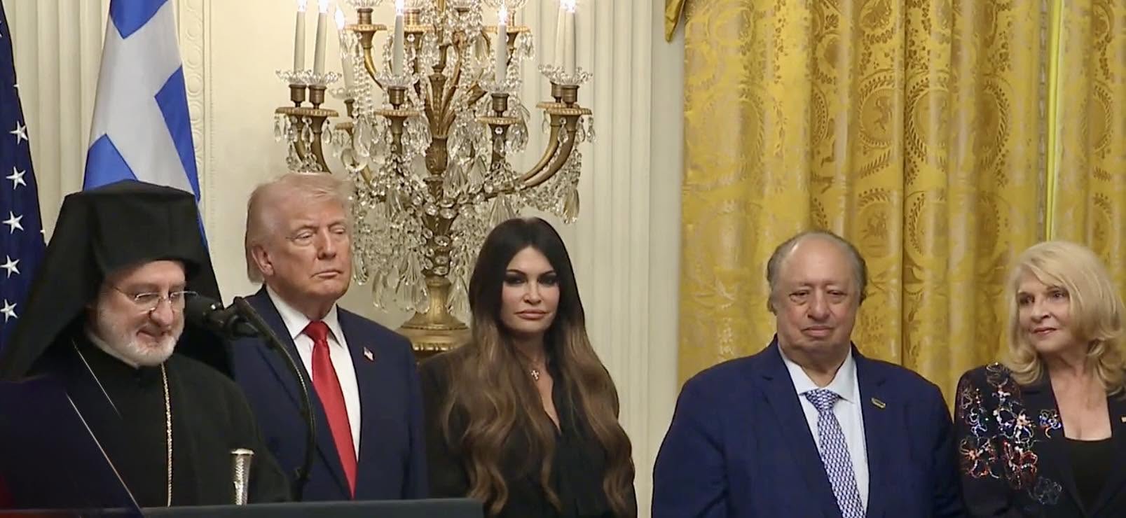 A video still of Greek Independence Day at the White House, with President Donald Trump, an Orthodox priest, and guests beneath a crystal chandelier.