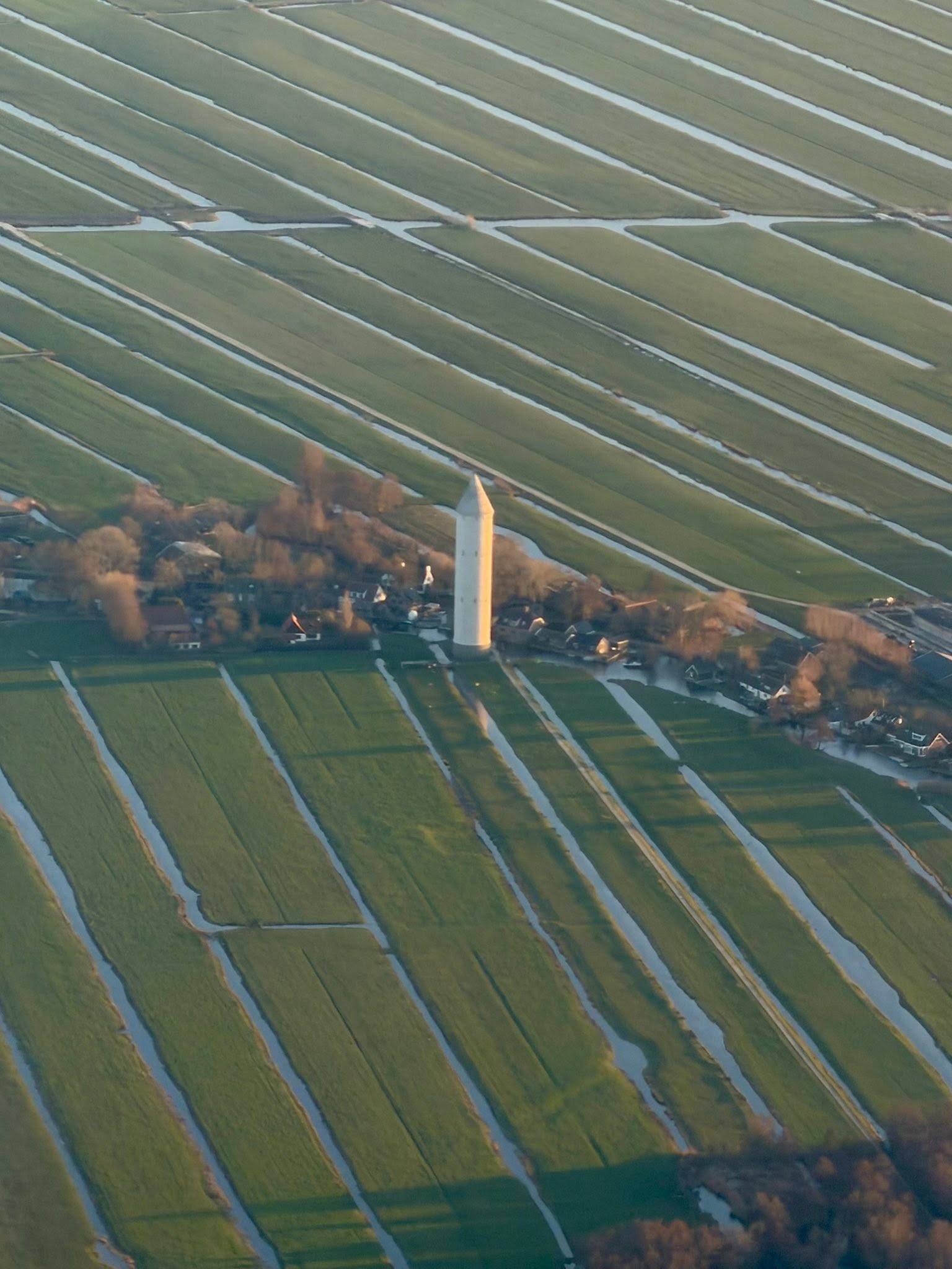 An aerial view of the Dutch polder landscape — green fields striped with water channels, a white church tower rising from a village at the center.