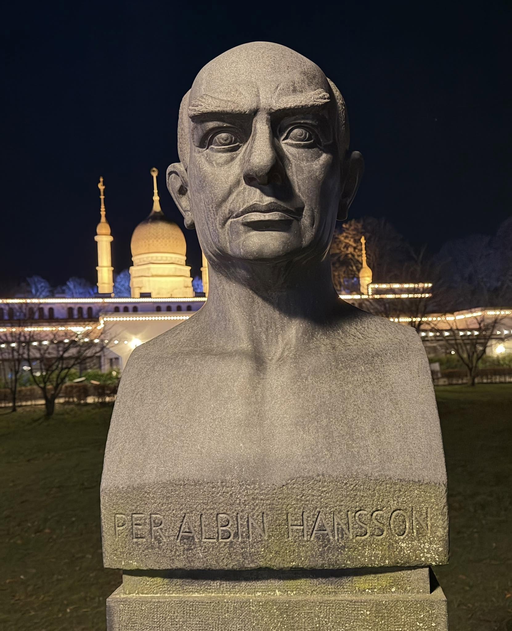 The bust of Per Albin Hansson photographed at night, an illuminated domed building glowing behind it.