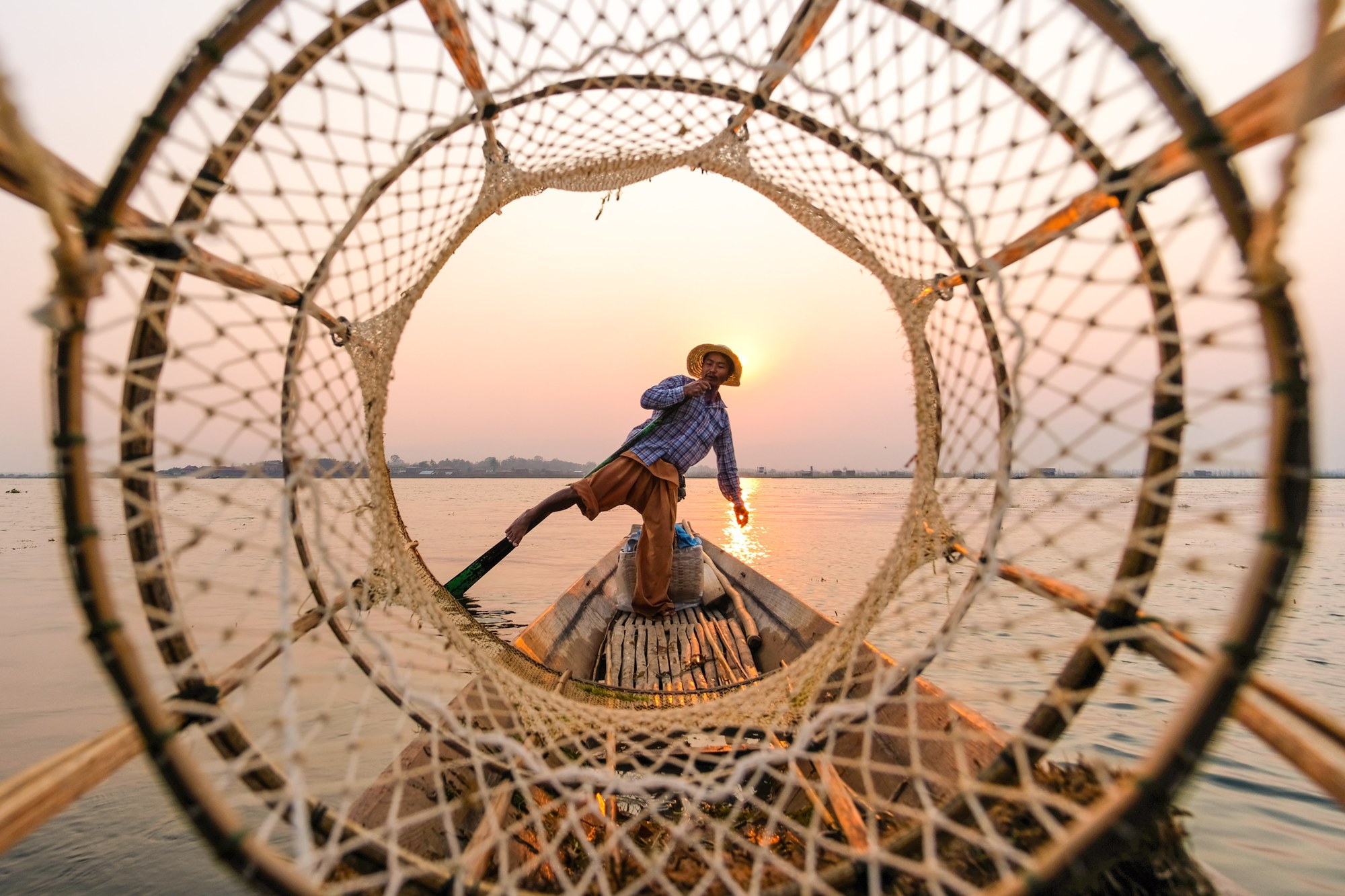 An Intha fisherman on Inle Lake balancing on one leg at the stern of his boat, framed through a conical fishing basket at sunset