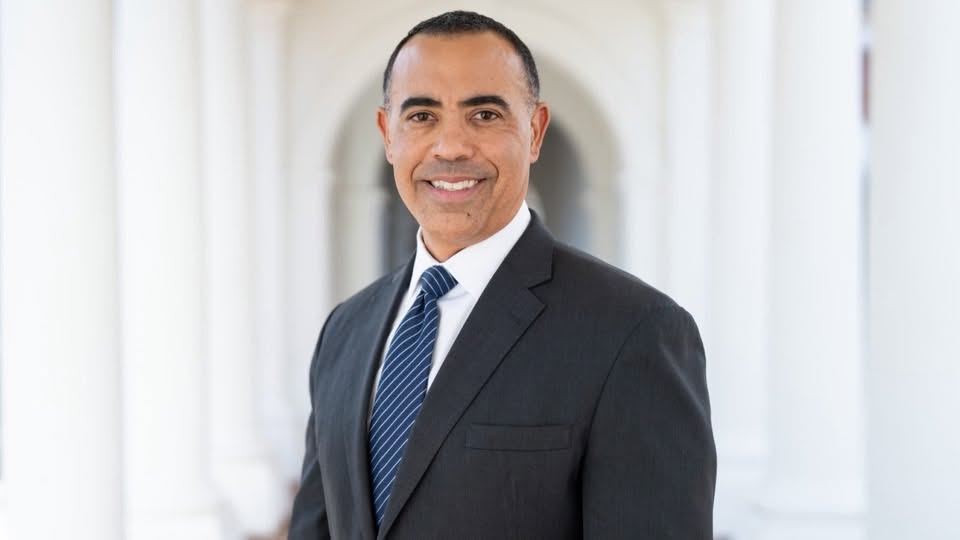 Ian Solomon, smiling, in a dark suit and striped tie, photographed before a white colonnade.