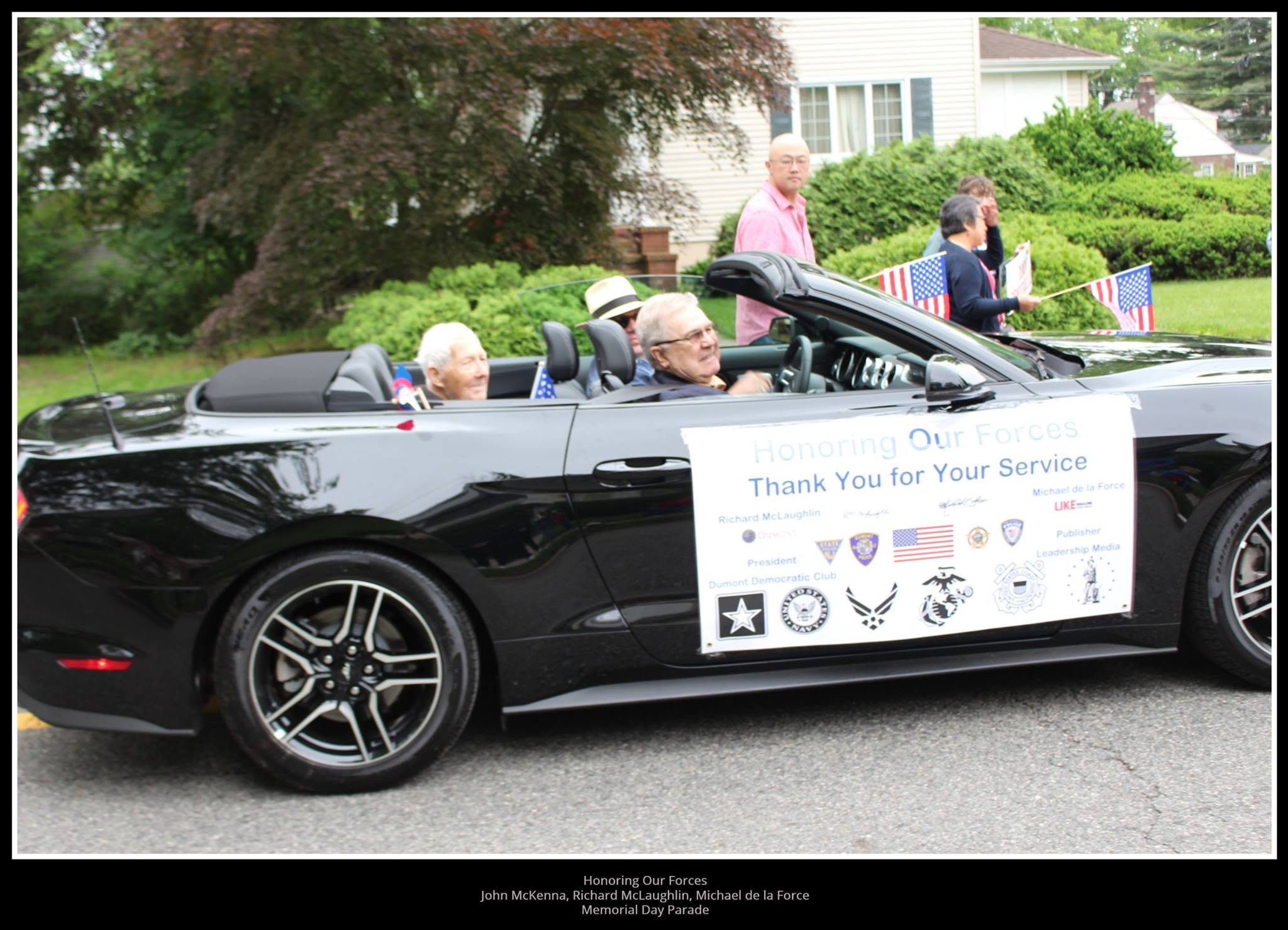 A black convertible Mustang carrying a 'Honoring Our Forces — Thank You for Your Service' banner in a Memorial Day parade.