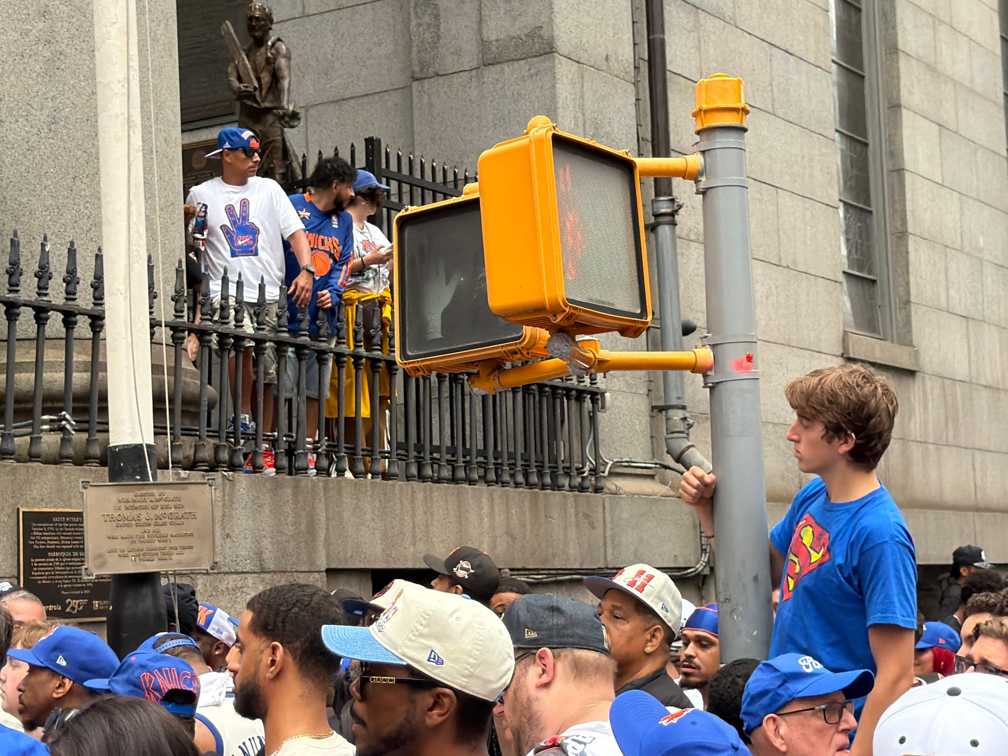 Knicks fans stand on the stone base behind the iron fence of Old St. Peter's Church beside a statue and a memorial plaque, while a boy in a blue shirt holds a crosswalk signal pole above a crowd in Knicks caps