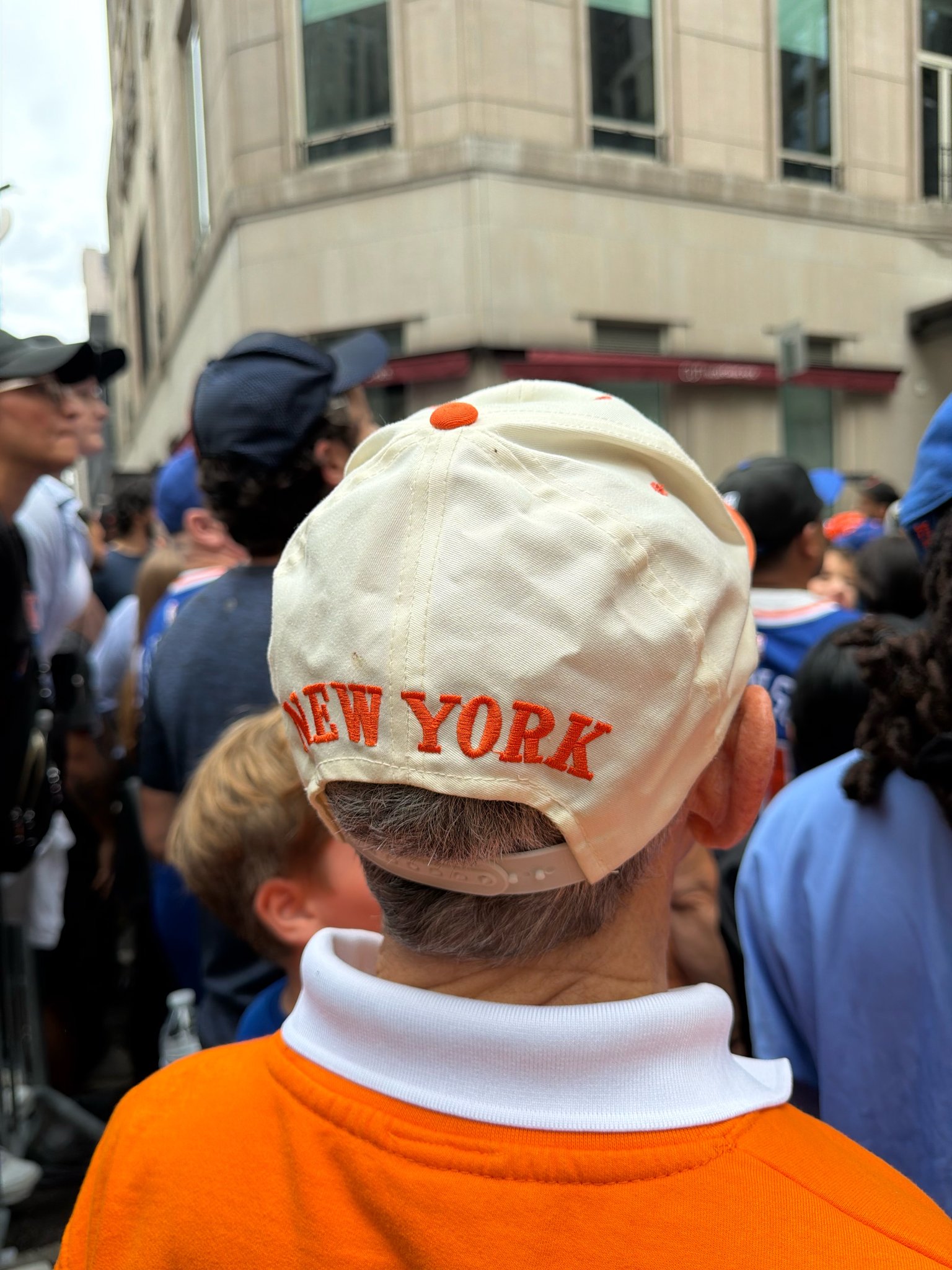 The back of a cream baseball cap embroidered NEW YORK in orange, worn with an orange shirt, in the parade crowd