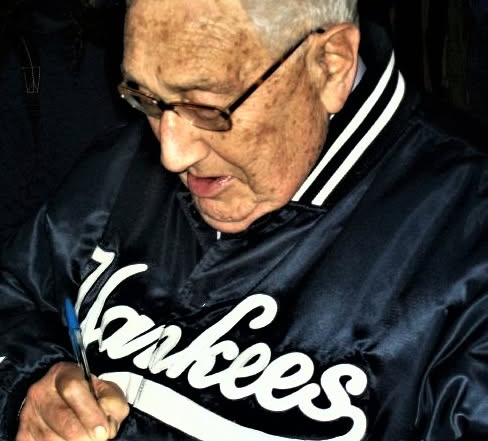 Henry Kissinger, in glasses and a navy Yankees satin jacket, writes with a blue pen