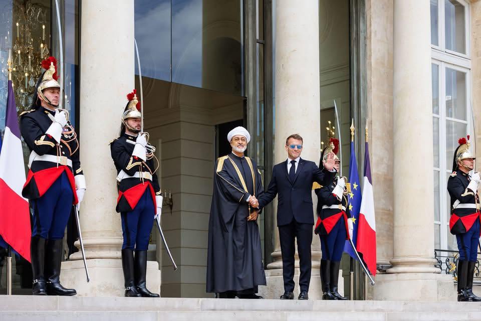 Sultan Haitham bin Tariq of Oman, in traditional dress, shakes hands with French President Emmanuel Macron on palace steps, flanked by Republican Guards in ceremonial uniform.