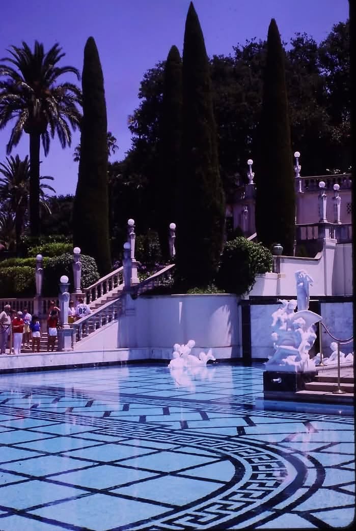 The Neptune Pool at Hearst Castle in San Simeon, California — blue-and-white geometric tilework, marble statues, tall cypress trees, and visitors on the grand staircase.