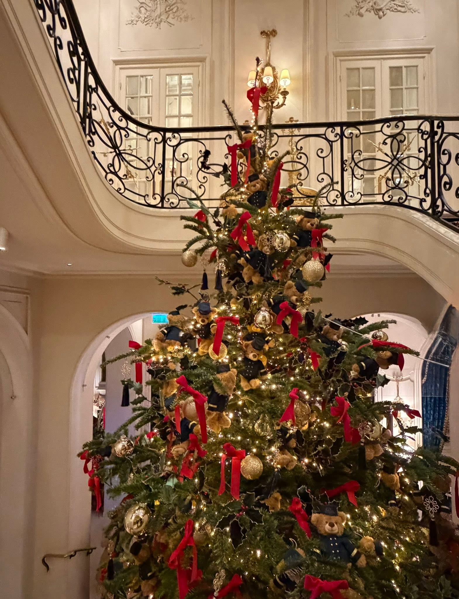 The Ritz's Christmas tree, trimmed with red bows, gold baubles, and teddy bears in uniform, rising beside the grand staircase.