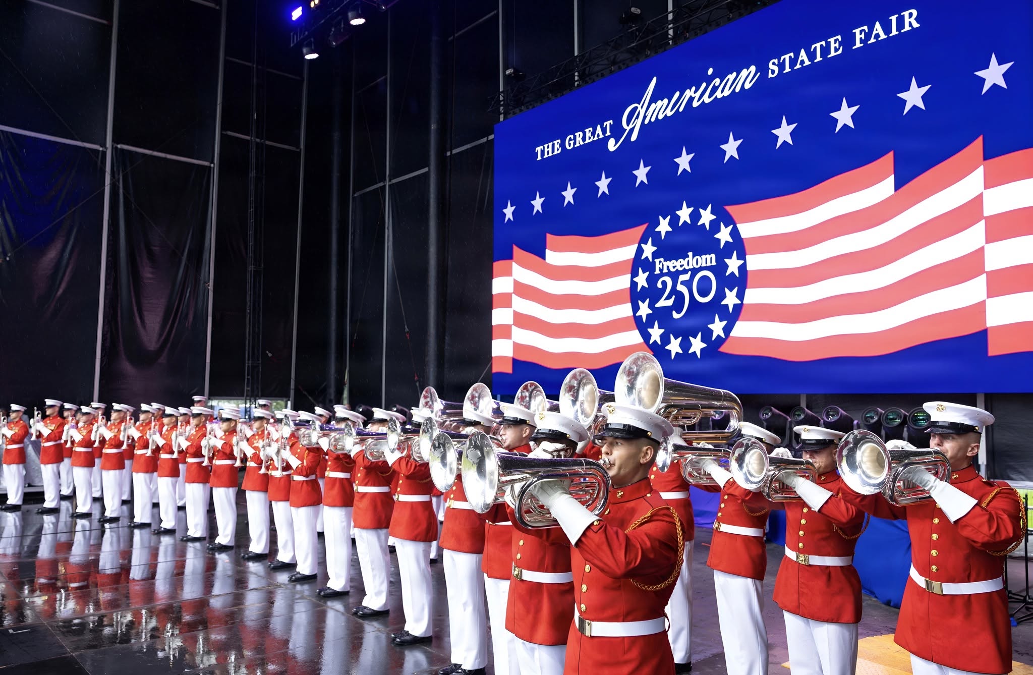 Members of the U.S. Marine Corps band in red dress uniforms perform on brass instruments onstage beneath a 'The Great American State Fair' and 'Freedom 250' backdrop.