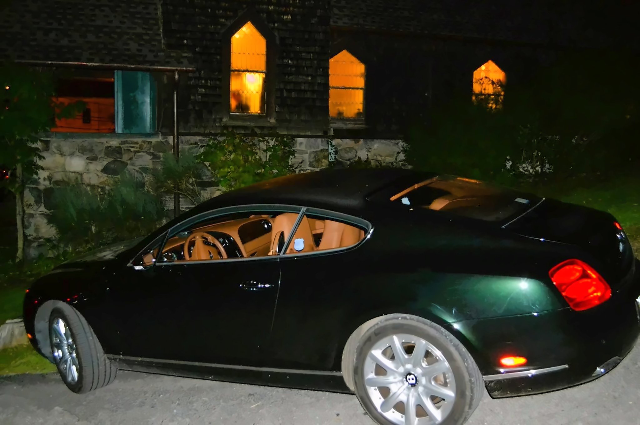 Night in Tuxedo Park: a dark green Bentley coupé, by its lines a Continental GT, with multi-spoke wheels, a winged-B center badge and a tan leather cabin, parked below lit pointed-arch windows.