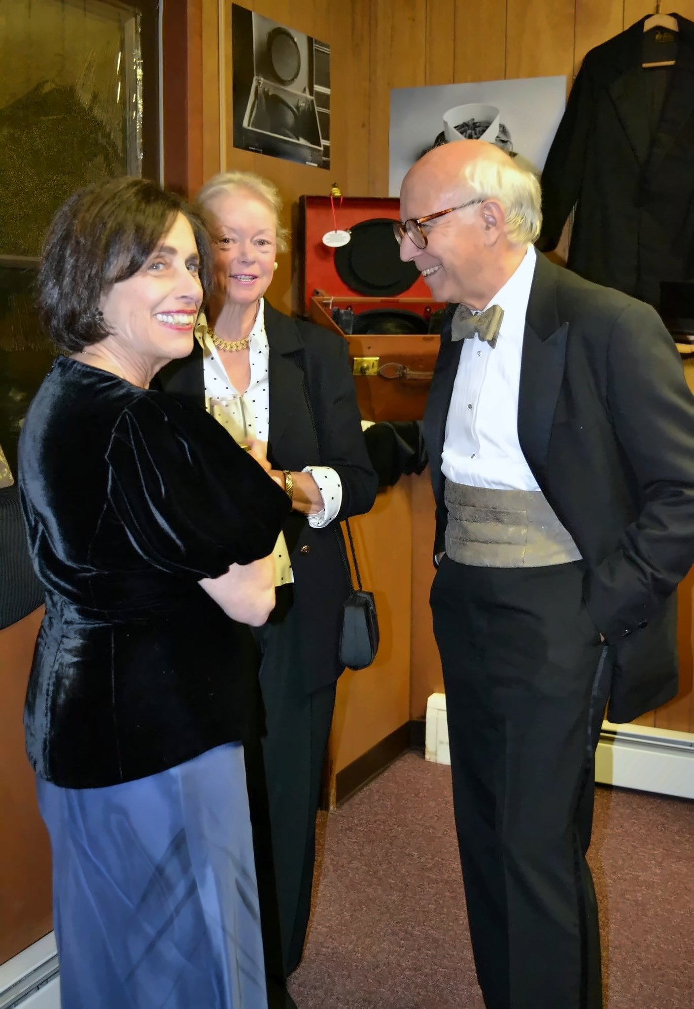 A pale silver-grey cummerbund under an open dinner jacket with a black bow tie; beside him, a black velvet jacket. Behind, the red-lined Victorian hat case and a tailcoat on display.