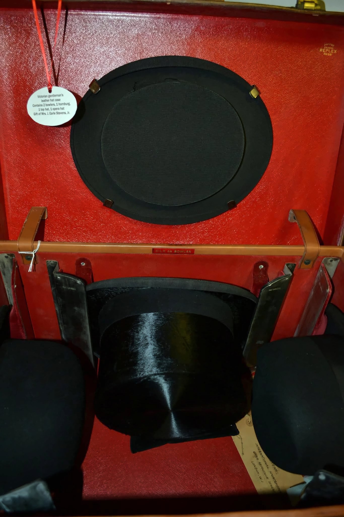 The case open: red leather, a round black lid compartment, and a top hat nested between the rounded crowns of hard hats.