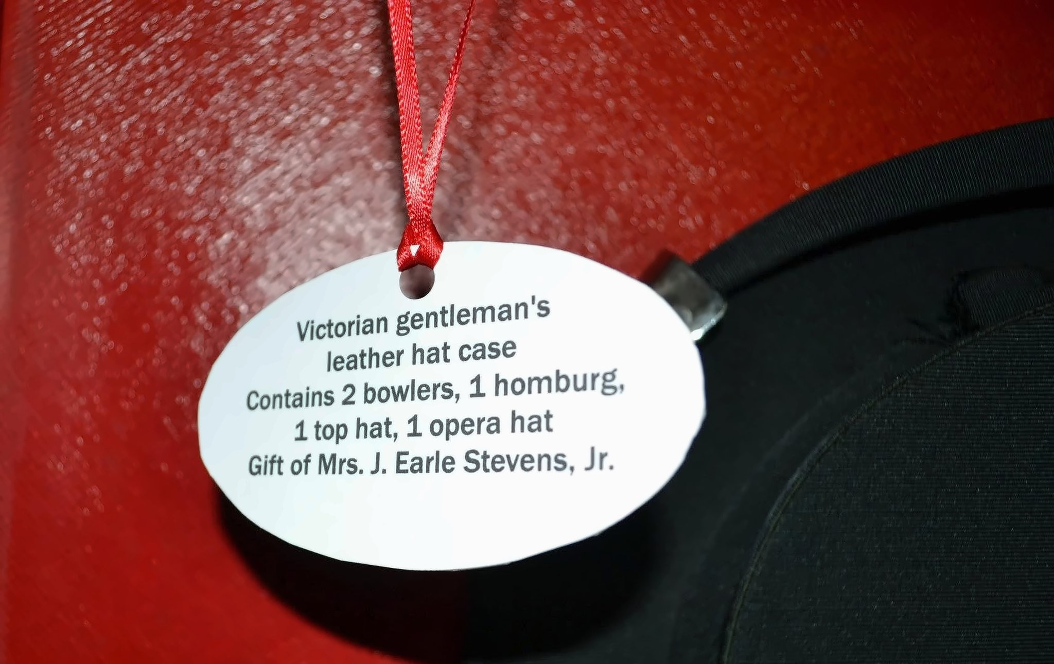 The oval tag: "Victorian gentleman's leather hat case. Contains 2 bowlers, 1 homburg, 1 top hat, 1 opera hat. Gift of Mrs. J. Earle Stevens, Jr." A black hat brim rests against the red leather interior.