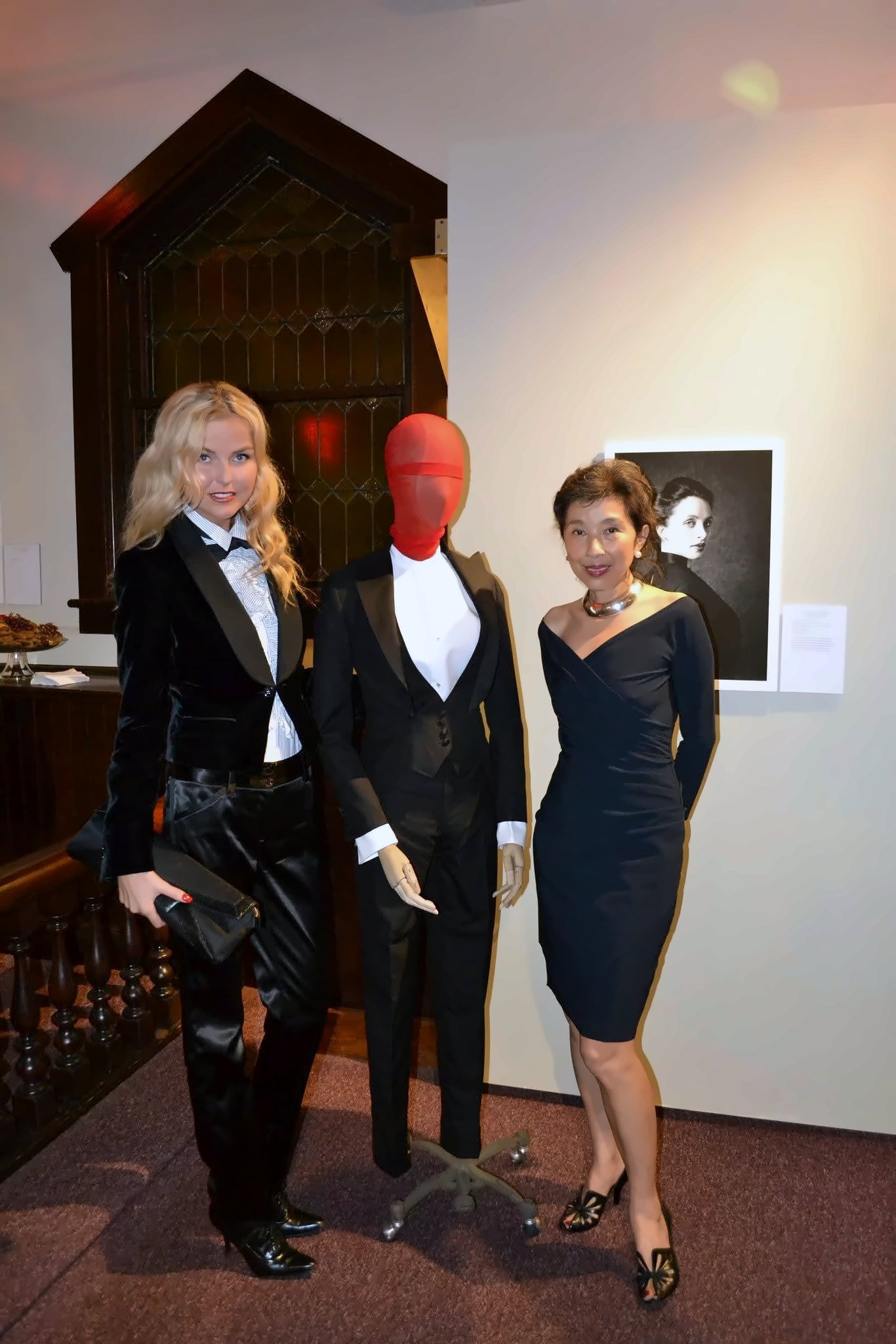 On either side of a red-faced mannequin in a black tuxedo: a black velvet tuxedo jacket over a white shirt and trousers, and a black off-the-shoulder cocktail dress with a statement necklace.
