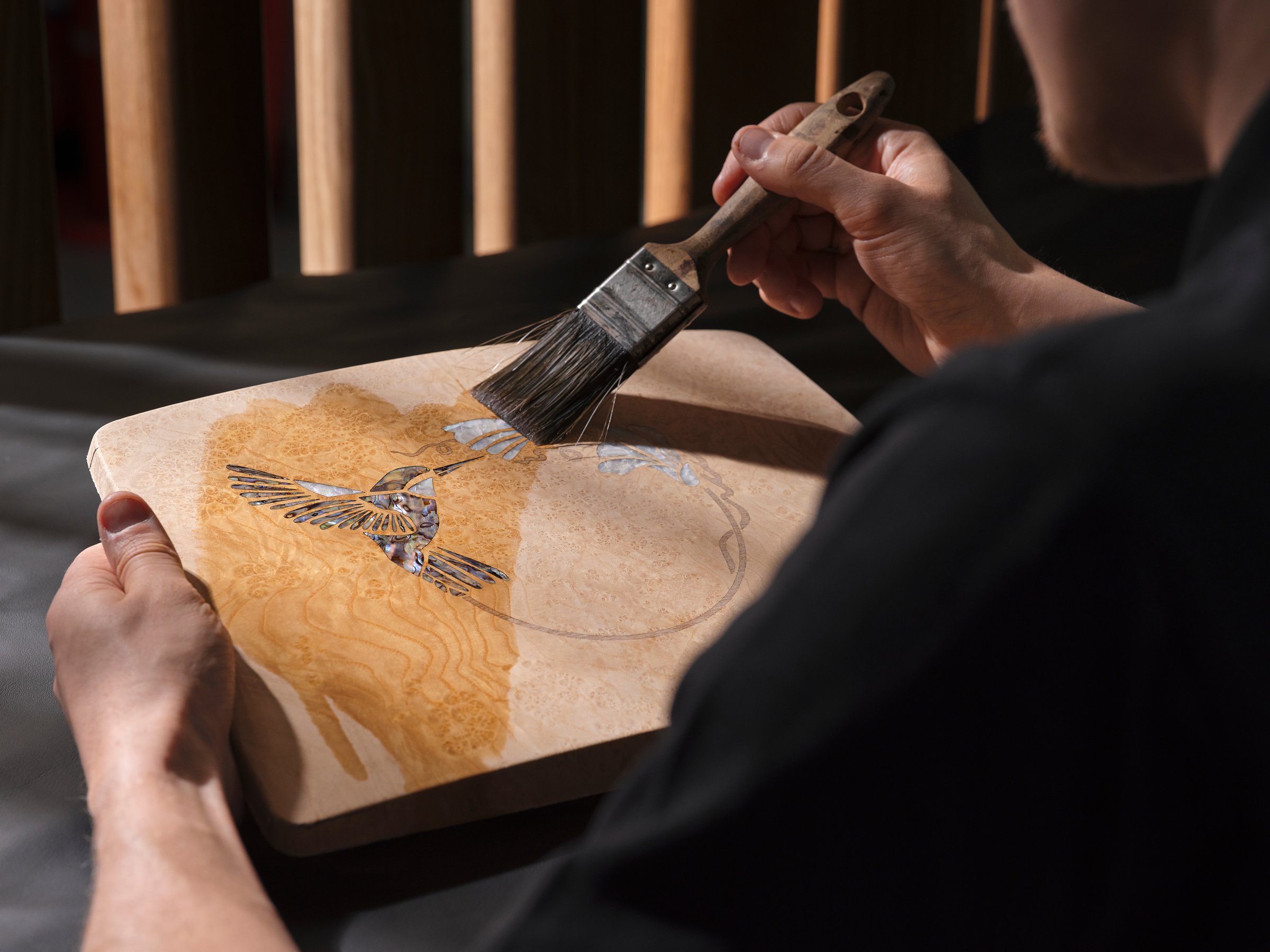 A craftsperson brushing lacquer over the hummingbird marquetry panel